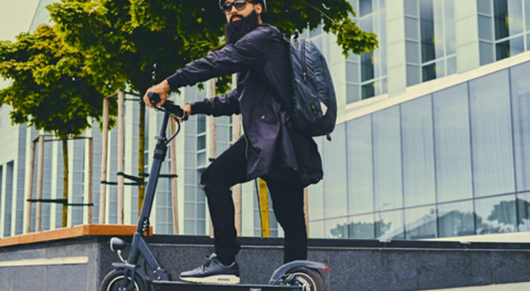 Stylish bearded male in sunglasses posing on electric scooter in over modern building background.
