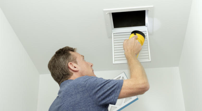 Man inspecting air ducts shining a flashlight through a small square ceiling vent into ducting pipes.