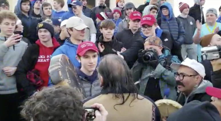 In this Friday, Jan. 18, 2019 image made from video provided by the Survival Media Agency, a teenager wearing a "Make America Great Again" hat, center left, stands in front of an elderly Native American singing and playing a drum in Washington. The Roman