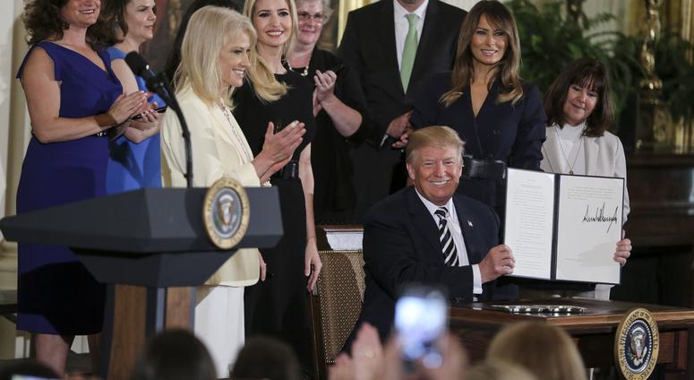 President Donald Trump, joined by Counselor to President Donald Trump Kellyanne Conway, Ivanka Trump, first lady Melania Trump, Karen Pence and others, holds up an executive order to address military spouse unemployment at the White House, May 9, 2018.