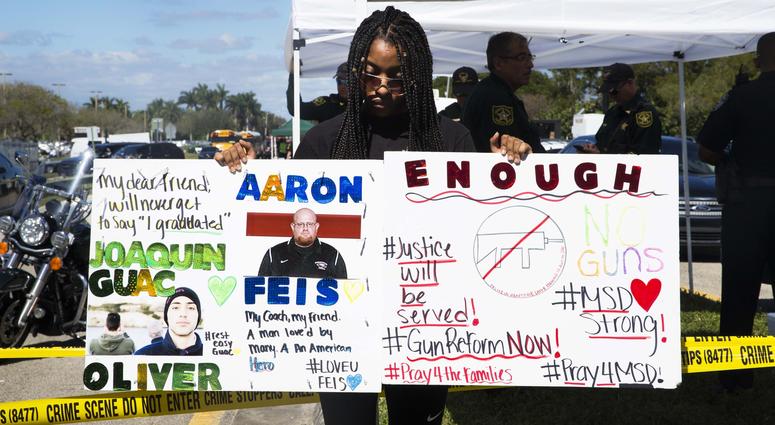 Tyra Hemats, 19, a 12th grader at Marjory Stoneman Douglas High School, stands with signs one advocating for gun reform and another honoring her coach and friend, both of whom were killed in Wednesday's shooting.