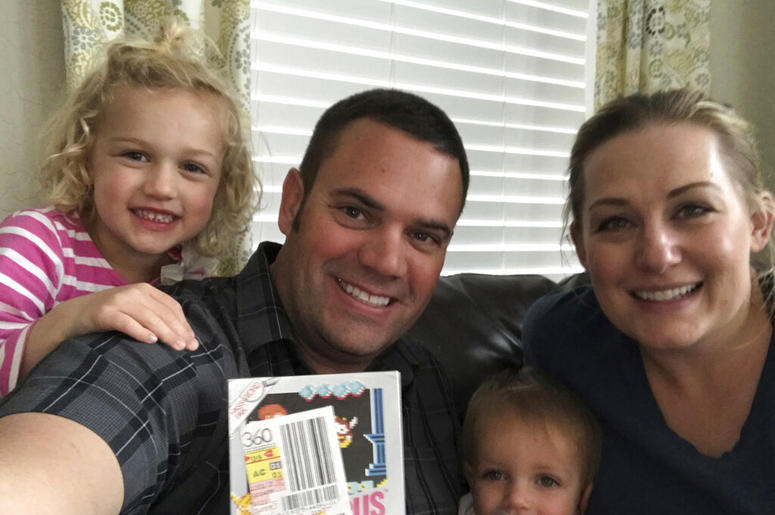 In this June 15, 2019 photo provided by Heritage Auctions, Scott Amos, along with his wife, Kristy, and daughters Grace, left, and Katie, pose in Reno, Nev., with an unopened copy of a 1987 cult-classic video game "Kid Icarus." The boxed game cartridge, s