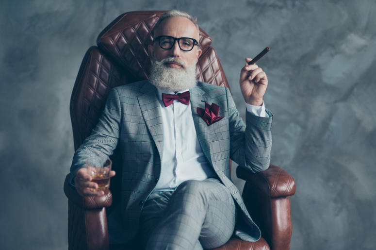 Cool man in glasses, hold cigarette, glass with brandy, in formal wear, tux with red bowtie and pocket square, sit in leather chair over gray background, looking to the camera, shares, stock, money