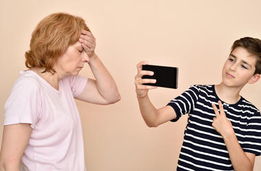 Youtuber Besa A Su Madre Por Fama Son taking selfie while his dissatisfied mother is slapping her head with hand. Family and modern technology addiction concept.