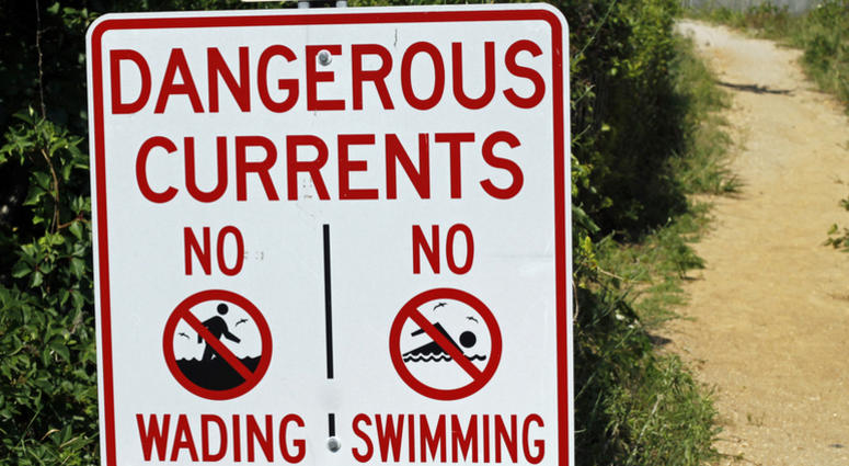 Coastal beach warning sign that says,Dangerous Currents, No Wading,No Swimming. Green shrubbery and sand path to beach. Swimming Warning