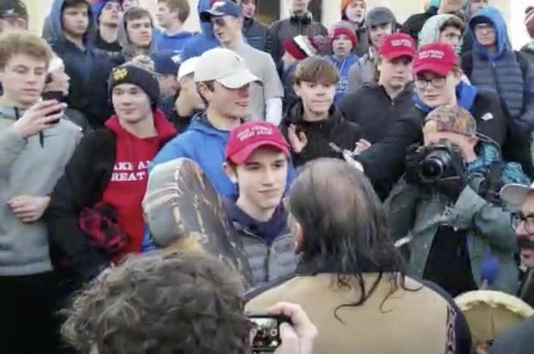 AP19019762910578 In this Friday, Jan. 18, 2019 image made from video provided by the Survival Media Agency, a teenager wearing a "Make America Great Again" hat, center left, stands in front of an elderly Native American singing and playing a drum in Washington. The Roman