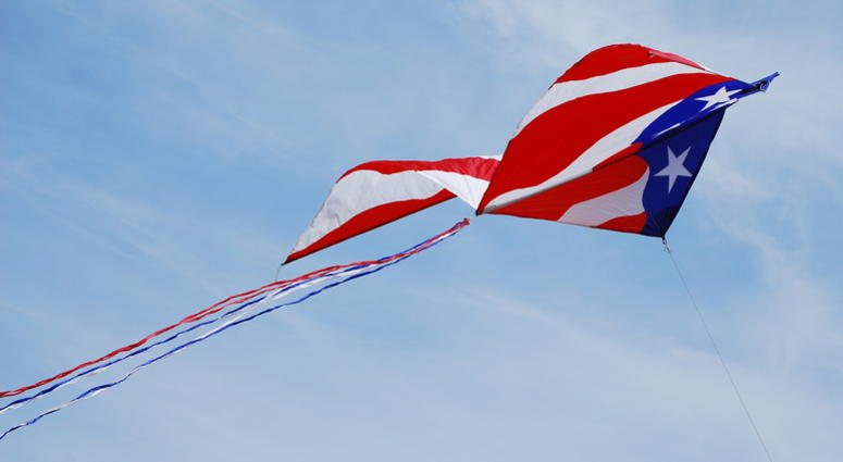 A kite with a Stars and Stripes theme, trailing red white and blue tail streamers soars diagonally across the frame, giving a sense of speed and movement. A kite with a Stars and Stripes theme, trailing red white and blue tail streamers soars diagonally across the frame, giving a sense of speed and movement.