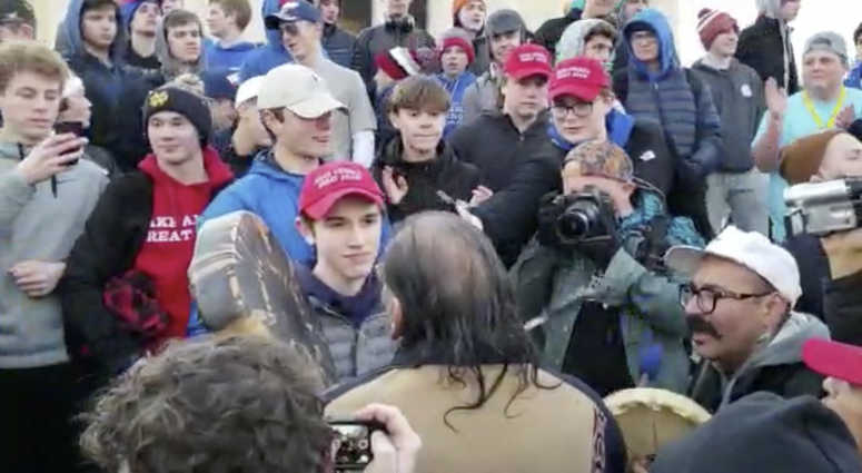 Media Agency, a teenager wearing a "Make America Great Again" hat, center left, stands in front of an elderly Native American singing and playing a drum in Washington. MAGA hat Native American Washington